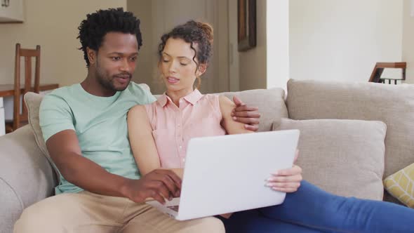 Happy biracial couple sitting on sofa with laptop and talking