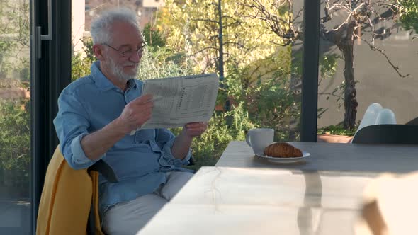Senior man reading newspaper at breakfast table in sunny kitchen