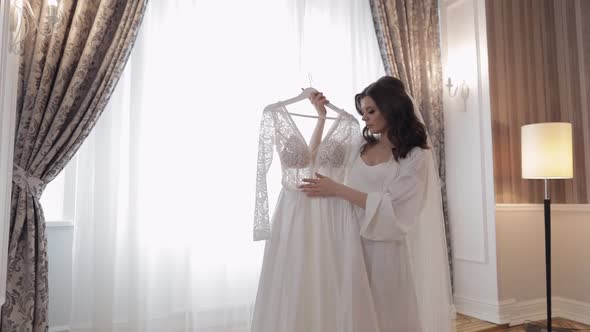 Bride in Lingerie Standing with Her Wedding Dress at Home Near Window White Boudoir Dress and Veil