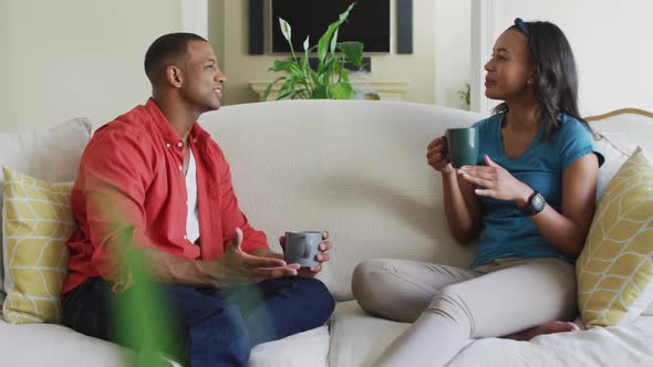 Happy biracial couple sitting on sofa with coffee and talking