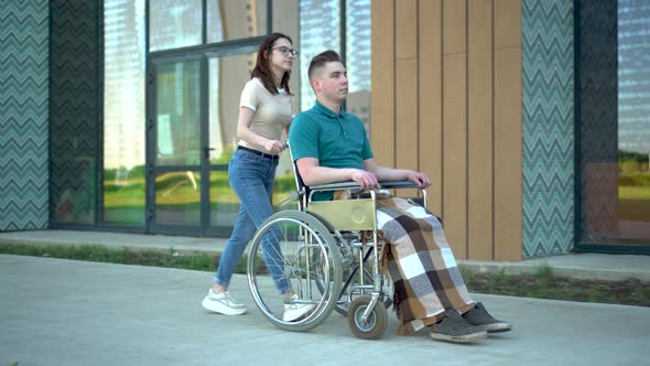 A Young Woman Is Carrying a Young Man in a Wheelchair. An Assistant Carries a Disabled Person in a