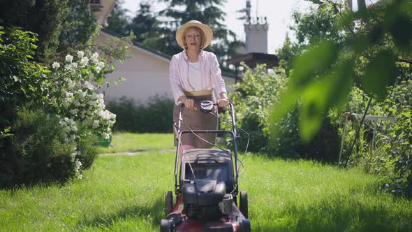 Positive Confident Senior Woman Walking in Sunshine Trimming Grass on Backyard with Lawn Mower in