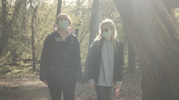 Middle Shot of Two Girls in Face Masks Strolling in Spring Forest and Leaving. Caucasian Women