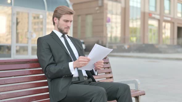 Young Businessman Reading Documents While Sitting on Bench Outdoor