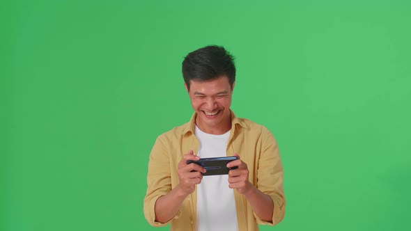 Laughing Asian Man Looking At The Phone Screen And Holding It In Her Hands In Green Screen Studio