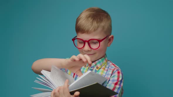 Elementary Age Boy in Eye Glasses Reading Book and Smiling Standing on Blue Background