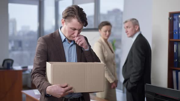 Portrait of Sad Fired Young Man Standing in Office with Box As Blurred Colleagues Talking at