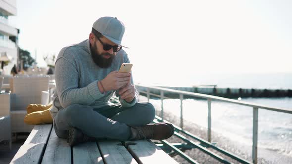 Young Man Is Using Internet on His Mobile Sitting on Wooden Boards Near Sea