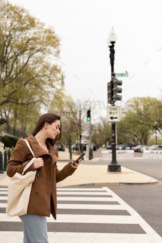 Young female is standing on a pedestrian crossing, observing the screen of her mobile device