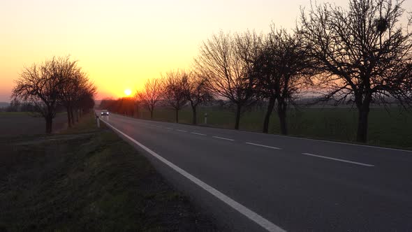 A Car Drives Up a Highway Through a Rural Area  the Golden Sunset in the Background
