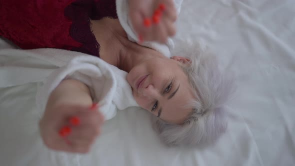 Smiling Mature Woman Stretching Hands to Camera Lying on White Comfortable Bedding