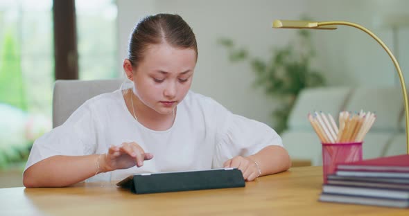 Focused Teen Girl at the Desk Scrolling Tablet Books Pencils and Lamp on the First Plan First Plan