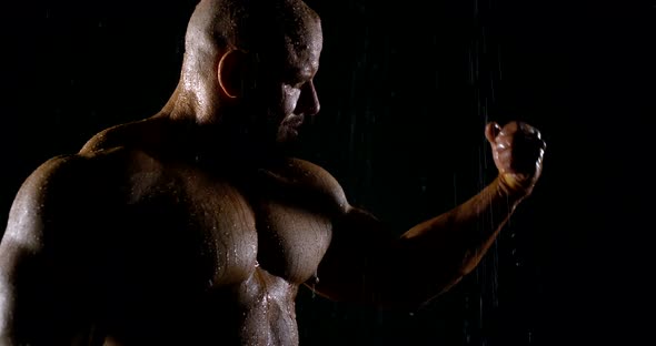 Bodybuilder Is Bending Hands and Showing Biceps and Triceps, Standing Under Rain in Night