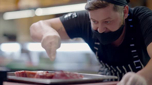 Confident Professional Male Cook in Covid Face Mask Salting Raw Meat on Baking Sheet in Slow Motion