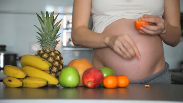 Pregnant Woman Peels a Tangerine in Home Kitchen with Different Tropical Fruits