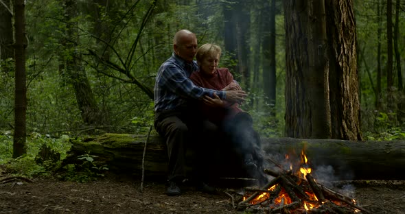 Senior Citizen in Shirt Hugs Wife Sitting at Fire in Forest