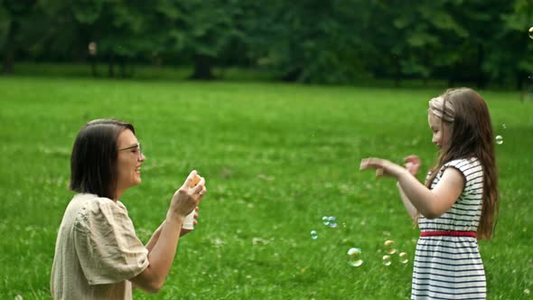 Beautiful Young Mother Together with Her Daughter in Nature Making Soap Bubbles and Laughing