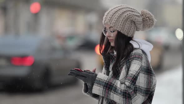 Closeup of a Young Darkhaired Woman in Round Glasses and a Knitted Hat is on a City Street in the