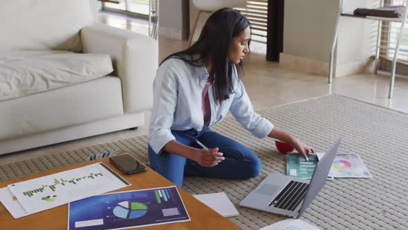 Mixed race woman sitting on the floor at home using laptop and writing in notebook