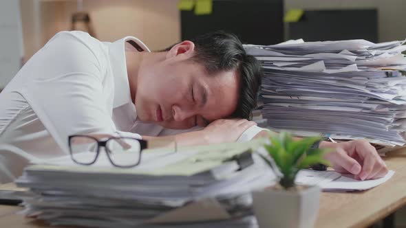 Close Up Of Tired Asian Man Sleeping Due To Working Hard With Documents At The Office