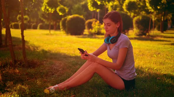Cheerful Female Listening Music Sits on the Green Grass