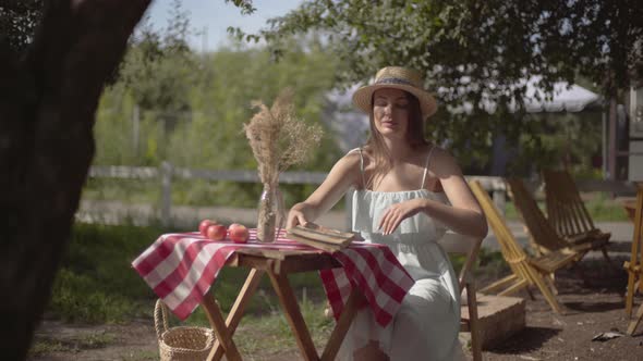 Young Girl in a Straw Hat and White Dress Finishing To Read the Old Book Sitting at the Small Table