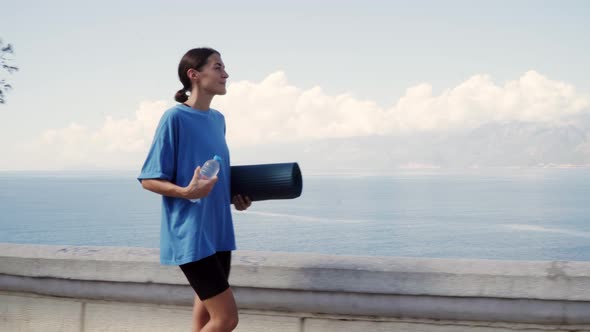 A Woman with a Yoga Mat and Water Walks Along the Sea for a Workout