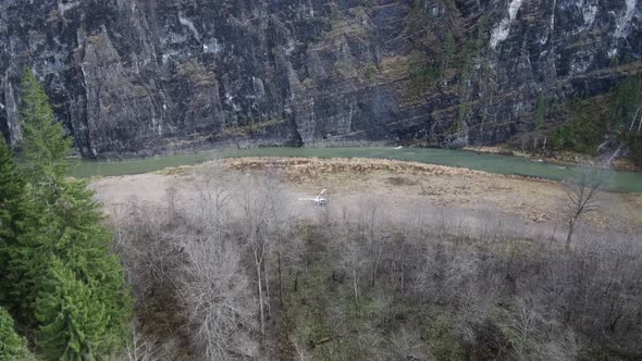 A Helicopter with Spinning Propellers Stands on the River Bank Under a Rock