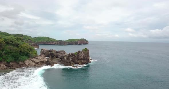 Aerial flyover coastline of Pantai during cloudy day with crashing waves against rocks on shore