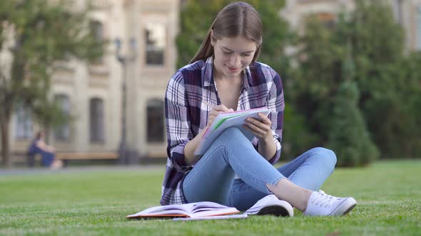 Student Sitting on The Green Grass and Writing in The Paper Notebook