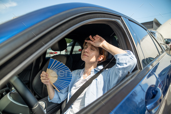 Exhausted middle aged woman drives car waves blue fan suffers from stuffiness in traffic jam.