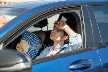 Exhausted middle aged woman drives car waves blue fan suffers from stuffiness in traffic jam.