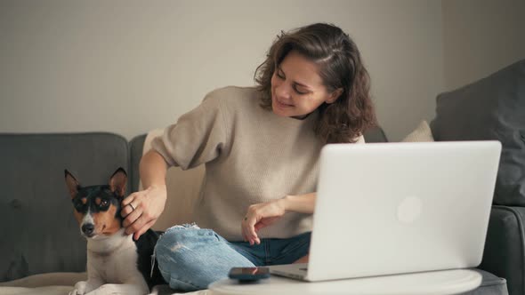 A Young Woman Working on a Laptop While Sitting on a Sofa with Her Cute Dog