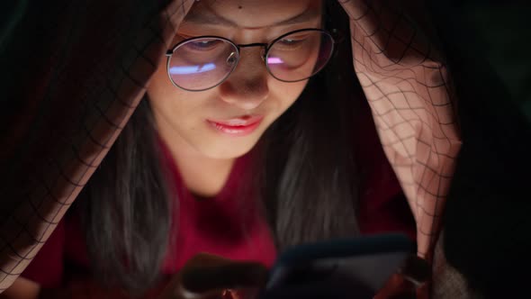 Close up young Asian woman using smartphone underneath duvet in the dark on bedroom at home.