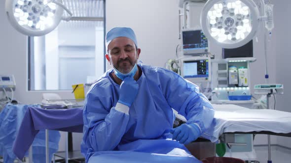 Portrait of caucasian male surgeon wearing lowered face mask sitting in operating theatre smiling