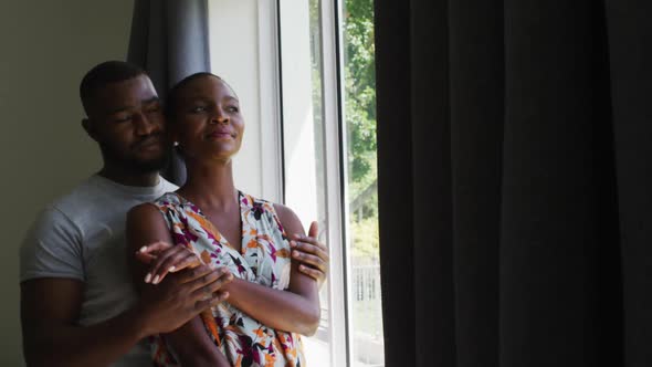 African american couple embracing each other while looking out of the window at home