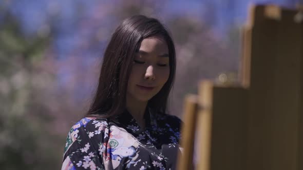 Talented Smiling Asian Woman Painting on Easel Standing in Sunny Sakura Garden and Drinking Tea