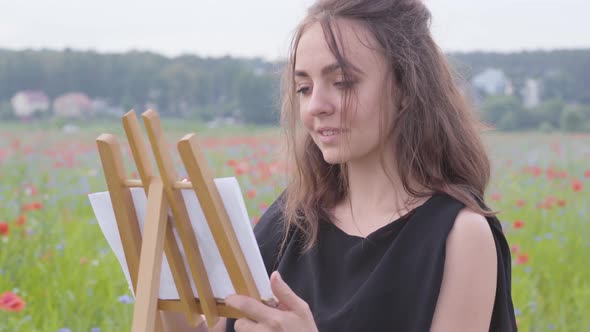 Portrait of Cute Smilling Girl Painting on the Easel Tanding on the Poppy Field. Young Woman Artist