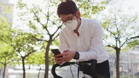 Asian man wearing face mask using smartphone while leaning on his bicycle on the street