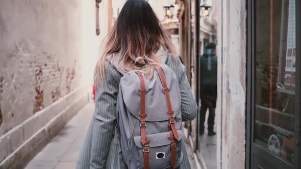 Camera Follows Stylish Female Tourist with Backpack and Long Hair Walking Along Old Summer Street in