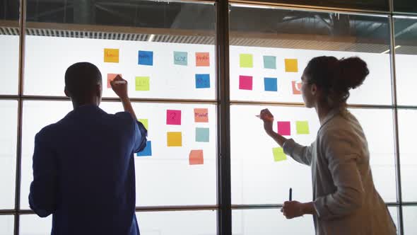 Diverse female creative businesswomen writing on memo notes, brainstorming on glass wall in office