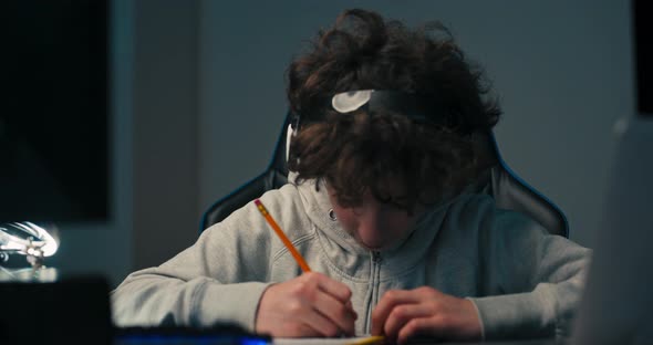 A Boy Sits in Room in the Evening in Front of Computer Screen and Laptop Working on a School Project