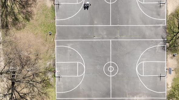 An aerial view over 4 people playing basketball on a court surrounded by dry trees on a sunny day. T