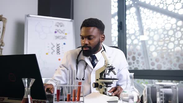 Medical Assistant which Looking Into Microscope and Writing Down the Results on the Laptop