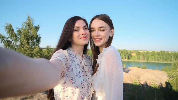 Two Pretty Young Passionate Women in Dresses in Mountains with Blue Lake in Background Make Selfie