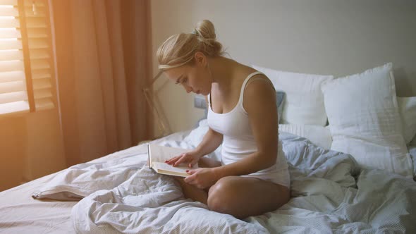 Lovely Young Woman in White Tank Top and Panties Sitting on Comfortable Bed and Reading