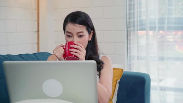 Asian woman working on laptop checking social media and drinking coffee while lying on the sofa.