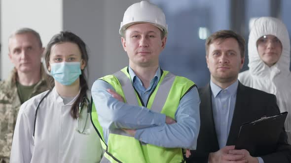 Confident Smiling Male Builder Looking at Camera Crossing Hands Standing Indoors with Group of