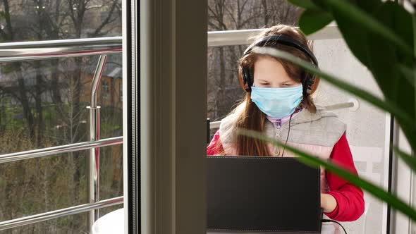 Teenager Girl in Mask and Headphones, Typing, Working on Laptop, on Open Balcony. Spring Sunny Day