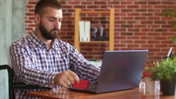 Disabled Hipster is Sitting in Wheelchair Holding Credit Card and Using Laptop Side View